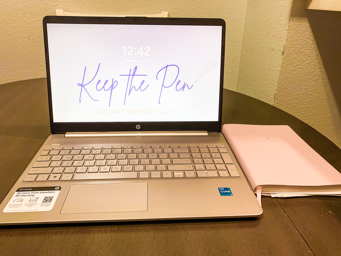 laptop sitting on a table and open to the home screen showing Keep the Pen Notary Services, LLC logo. A pink notebook sits beside the laptop