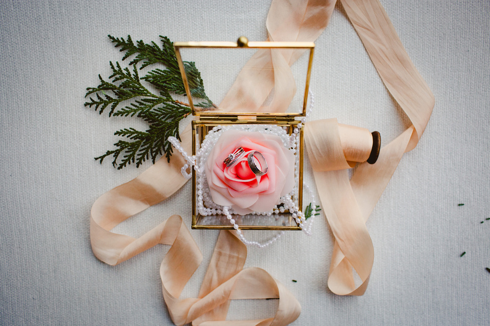 pink and white rose inside a glass box with gold trim. A string of pearls lays over it. Two wedding rings sit on top of the rose. champagne colored ribbon around the outside of the box. greenery is seen at the upper left corner of the box.